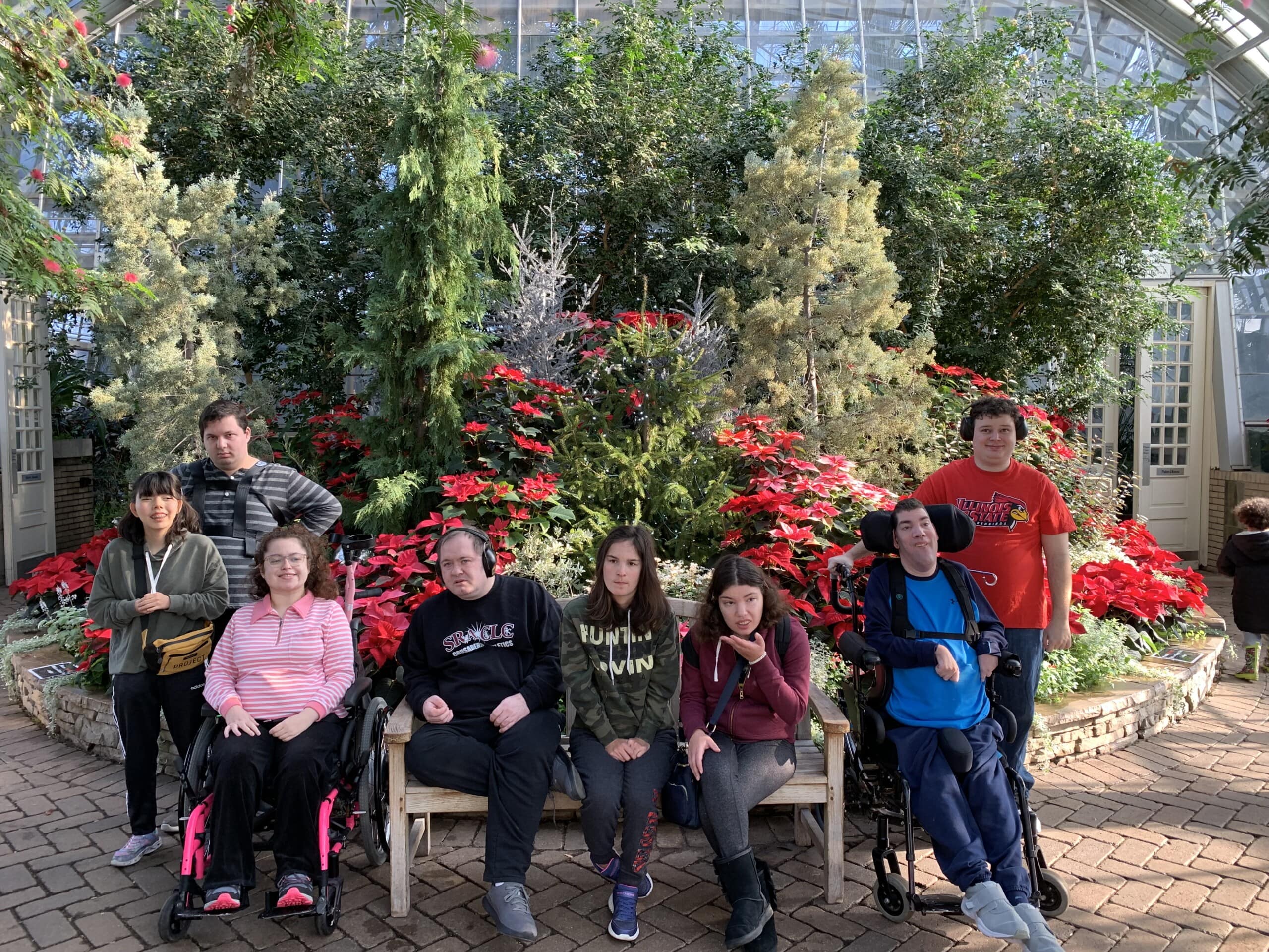 A group of CEL REACH members smiling and posing in the Garfield Park Conservatory greenhouse, with some members sitting on a bench and others standing.