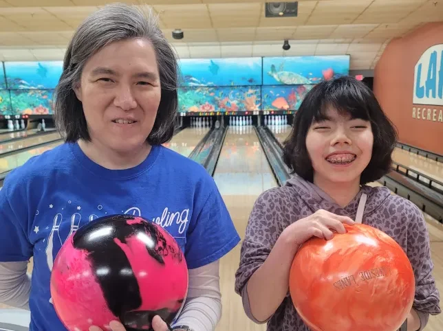 A member of CEL, smiling, holds bowling balls while standing next to a friend during Monday Night Bowling.